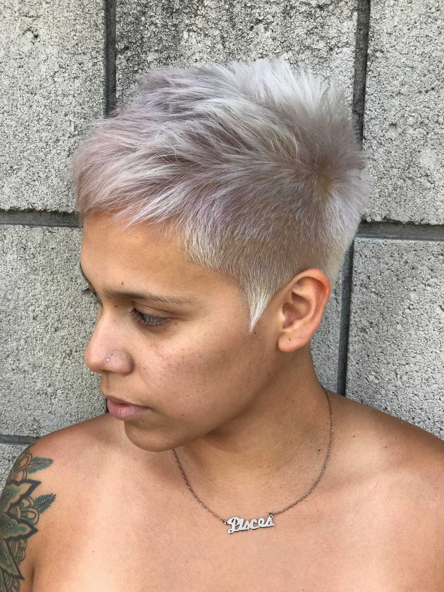 Person with short, spiky platinum blonde hair stands against a gray brick wall.