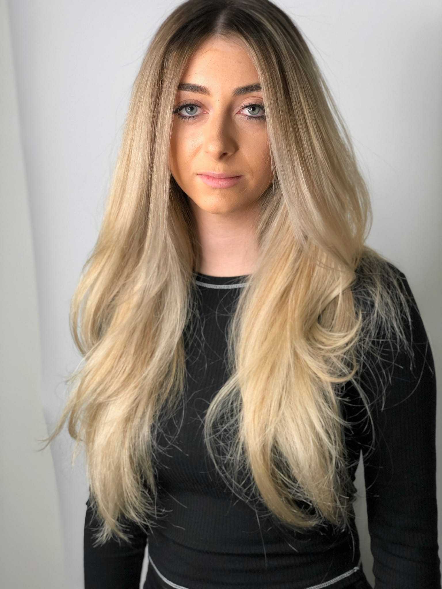 Woman with long blonde hair, wearing a black top, standing against a plain background.